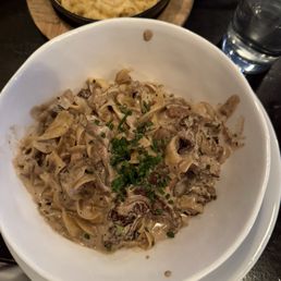 Beef Stroganoff - A bowl of beef stroganoff with creamy sauce and chopped herbs, accompanied by macaroni and cheese and a glass of water, on a dark table.