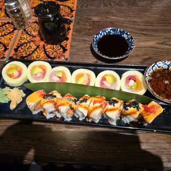 The 698 - Naruto Roll (back) White Dragon Roll (front) - The image shows a black rectangular plate with sushi rolls topped with sauces, black and red fish roe, and garnishes, placed on a wooden table. There are six sushi pieces, with a green leaf underneath, and condiments such as wasabi and pickled ginger on the side. Two small bowls containing soy sauce and a dark dipping sauce are positioned above the plate. The background includes a patterned tile surface, a glass of water, a black mug, and salt and pepper shakers. - Sushi Bars Near Me - Columbus, Ohio
