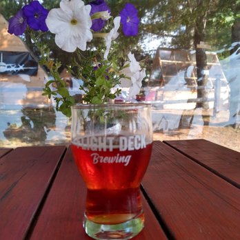 Flight Deck Brewing - Sorry y'all, couldn't resist the 1st Belgian beer on their rather elusive beer spreadsheet - A glass of amber-colored beer is placed on a wooden table, with a flower arrangement of white and purple petunias behind it. The scene is viewed through a window, reflecting trees and outdoor elements, indicating an outdoor seating area at Flight Deck Brewing. The glass has the brewery's logo on it. - Breweries Near Me - Brunswick, Maine