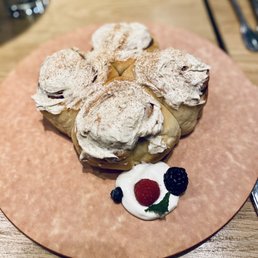 Cinnamon Roll - A plate features four cinnamon rolls topped with whipped cream and a dusting of cinnamon. The rolls are arranged on a round, pinkish plate placed on a wooden table. A dollop of whipped cream with a raspberry, blackberry, and a small blueberry is positioned beside the rolls, garnished with a small green leaf. The background includes utensils and a blurred setting, indicating a dining environment.