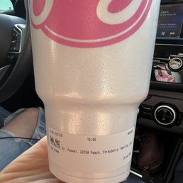 Boston - A large Styrofoam cup featuring a pink logo is held by a person's hand inside a car. The cup has a printed receipt at the bottom indicating a beverage order of an extra peach, strawberry, vanilla, and cream drink. The background shows part of the car's interior, including the dashboard, a radio display, and a person wearing ripped jeans. The scene suggests the person is in transit, possibly after purchasing a beverage.