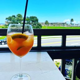 Sangria - A glass of sangria with lemon slices and a straw is positioned on a white table near a balcony railing. The background features a scenic outdoor view with green trees, a road with a vehicle, and a bright blue sky with some clouds. The setting suggests a relaxed dining or drinking experience outdoors.