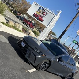 A gray sedan is parked in a parking lot near a sign with images of cars and an American flag, under a clear blue sky.