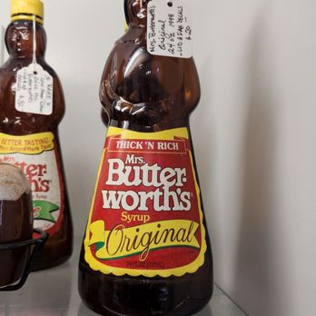 Blackberry Patch Antiques - A Mrs. Butterworth's syrup bottle with a yellow cap is placed on a glass shelf. The bottle has a label with red, yellow, and white colors, displaying the brand name and the word 'Original'. There is a handwritten tag attached to the bottle. The background is a plain white wall, and part of another similar syrup bottle is visible to the left. - Antiques Near Me - Powell, Ohio