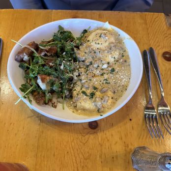 M'tucci's Italian - Biscuits and gravy - A white bowl on a wooden table holds biscuits covered in gravy with herbs and cheese, alongside a side salad of greens and grated cheese. Silverware is placed to the right of the bowl, and a salt shaker is visible at the bottom right. A person wearing a blue shirt is partially visible in the background. - Italian Near Me - Albuquerque, New Mexico