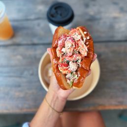 Lobster Roll - A lobster roll with seafood salad is held in a person's hand over a wooden table, with a coffee cup and a drink in the background.