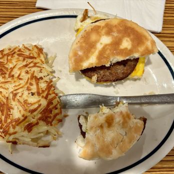 IHOP - A breakfast plate includes a burger with a bun, a beef patty, and a fried egg, along with hash browns and a biscuit. The plate is placed on a wooden table, with a metal utensil resting on the biscuit. A napkin is visible at the top of the image. - Breakfast & Brunch Near Me - Clinton, Maryland