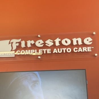 Firestone Complete Auto Care - They're awesome! - The image shows a sign for Firestone Complete Auto Care mounted on a reddish wall. The sign features the Firestone logo and text in white, with the phrase 'Complete Auto Care.' Part of a television screen is visible below the sign, displaying a person's face in a close-up shot. The setting appears to be inside a Firestone auto service center. - Tires Near Me - Firestone, Colorado