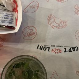 A glass containing a beverage with ice cubes and herbs is positioned on a seafood-themed tablecloth decorated with illustrations of lobsters, crabs, and other seafood items. The tablecloth also displays the restaurant's logo and branding for Cap't Loui, a seafood restaurant known for seafood boils. A paper bag with condiments or utensils is partially visible in the upper left corner.