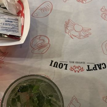 Cap't Loui - A glass containing a beverage with ice cubes and herbs is positioned on a seafood-themed tablecloth decorated with illustrations of lobsters, crabs, and other seafood items. The tablecloth also displays the restaurant's logo and branding for Cap't Loui, a seafood restaurant known for seafood boils. A paper bag with condiments or utensils is partially visible in the upper left corner. - Cajun/Creole Near Me - Providence, Rhode Island