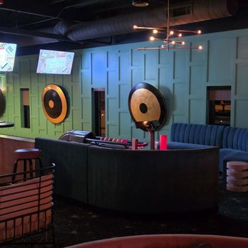 Palma - The image shows a lounge or bar interior with a green paneled wall decorated with large circular wall art pieces. There is a curved seating area with a blue velvet sofa and small tables. Multiple television screens are mounted on the wall, displaying colorful graphics. The space has low lighting, modern light fixtures, and a dark ceiling with exposed ductwork. Furniture includes chairs with cushions and a pool table partially visible on the left. - Sushi Bars Near Me - Phoenix, Arizona