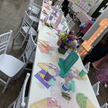 Play Modesto - A decorated party table features pastel-colored plates, cups, and utensils arranged for a children's celebration. The table is adorned with floral centerpieces, themed decorations, and a pink and purple color scheme. Additional party decorations include balloons, a large light-up letter, and a backdrop with balloons and banners. The setting appears to be indoors, with chairs arranged around the table and other party elements visible in the background. - Indoor Playcenter Near Me - Modesto, California