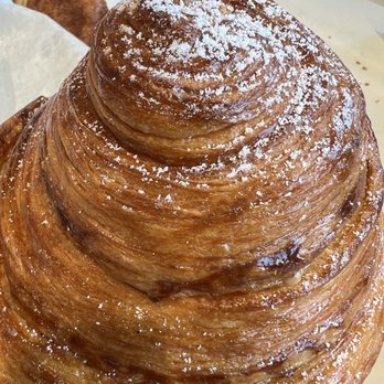 Tous Les Jours - A detailed view of a large, spiral-shaped croissant with a flaky, golden-brown crust. The pastry is dusted with powdered sugar on top and appears freshly baked. It is placed on a tray or plate, with part of another pastry visible in the background. The image emphasizes the layered, crispy texture of the croissant. - Coffee & Tea Near Me - Midvale, Utah
