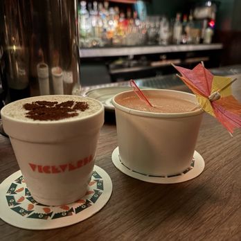 ViceVersa Miami - Two beverages are placed on a wooden bar counter, each in a white cup with different garnishes: one with a paper umbrella and the other with a small red stir stick. The drink on the left appears to be a creamy cocktail topped with cocoa powder, while the right drink is a pinkish cocktail with a decorative umbrella. The background shows a bar with shelves stocked with various bottles of alcohol and bar tools, creating a lively bar atmosphere. - Cocktail Bars Near Me - Miami, Florida