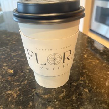 Flora Coffee - A paper coffee cup with a black plastic lid from Flora Coffee in Austin, Texas, sits on a dark granite countertop in a kitchen setting. - Coffee & Tea Near Me - Austin, Texas