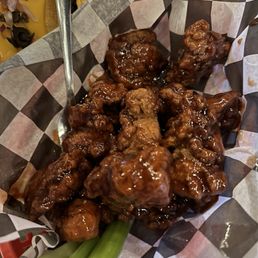 Boneless Wings - Basket of glazed chicken wings coated in a dark, sticky sauce, placed on checkered paper inside a basket. Green onion stalks are used as garnish at the bottom of the basket. The wings appear crispy and are arranged in a casual presentation, typical of bar or restaurant settings.