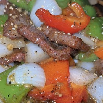 The 698 - Pepper steak - The image shows a close-up of a stir-fried dish with colorful bell peppers, onions, and beef strips, garnished with sesame seeds. The ingredients are served on a hot, black sizzling plate, highlighting the vibrant colors and textures of the vegetables and meat. - Sushi Bars Near Me - Columbus, Ohio