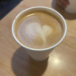 Plain latte with Pat milk - A disposable paper cup filled with a latte featuring foam art in the shape of a heart is positioned on a light wooden table. In the background, a person's hand holding a white cup is partially visible, with a blurred indoor setting. The foam art is centered on the coffee surface, and the shadow of the cup is cast on the table.