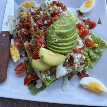 Ithaca Ale House - Salad - A salad on a white square plate includes sliced avocado, cherry tomatoes, chopped hard-boiled eggs, bacon bits, and shredded lettuce. The salad is topped with a creamy dressing and is presented outdoors on a wooden table. A small knife with a wooden handle is placed on the side of the plate. - New American Near Me - Ithaca, New York