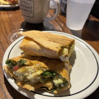 Corner Bakery Cafe - Avocado & Spinach Breakfast Wrap (V) - A plate holds a sliced breakfast burrito filled with eggs, vegetables, and cheese, with a toasted wrap placed on top. In the background, there is a mug of coffee and a plastic cup of water on a wooden table. The setting appears to be a casual dining environment, likely a cafe or restaurant. - Cafes Near Me - El Paso, Texas