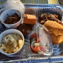 a tray of food - A disposable foil tray holds various food items including a serving of pasta with cheese, a sandwich bun, a small container of salad with sliced vegetables, and cooked meat. There are also small containers of condiments and a pen placed on the tray. The tray is set on a yellow checkered tablecloth, indicating outdoor dining or takeout service.