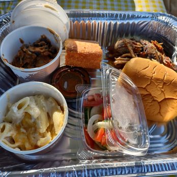 Moro's Kitchen - a tray of food - A disposable foil tray holds various food items including a serving of pasta with cheese, a sandwich bun, a small container of salad with sliced vegetables, and cooked meat. There are also small containers of condiments and a pen placed on the tray. The tray is set on a yellow checkered tablecloth, indicating outdoor dining or takeout service. - Italian Near Me - Skaneateles, New York