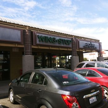 Wingstop - cars parked in front of a wingtop store - The image shows the exterior of Wing Stop, a restaurant specializing in wings, with a prominent sign displaying the business name. The storefront has large windows and an entrance door with an 'Open' sign. Several cars are parked in front of the restaurant, including a black Chevrolet with a visible license plate, and other vehicles in the parking lot. The sky above is partly cloudy, and the overall scene depicts a typical commercial area. - Chicken Wings Near Me - Phoenix, Arizona