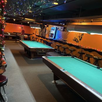 Atomic Billiards - The image shows a bar interior with two pool tables with green felt, positioned parallel to each other. The bar has orange walls decorated with a silhouette mural and string lights along the wall. Bar stools are lined up along the wall, and a vending machine is mounted on the wall. Overhead, colorful string lights hang from the ceiling, creating a festive atmosphere. A person is seen in the background near the bar area. - Pool Halls Near Me - Washington, DC, District of Columbia