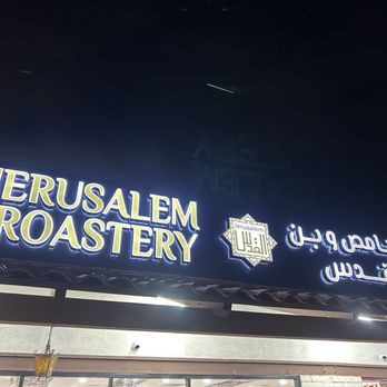 Jerusalem Roastery - The image shows an illuminated sign for Jerusalem Roastery, with the business name in large, bright letters in English and Arabic. The sign is mounted above the storefront, which is partially visible at the bottom of the image. The background features a dark night sky with a few faint stars or aircraft lights. The sign's lighting makes it clearly visible against the night. - Candy Stores Near Me - Garden Grove, California