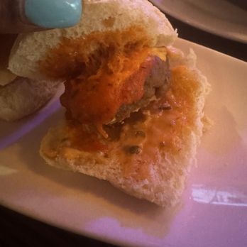 Patrick Steakhouse - Lamb Sliders (3) - The image depicts a beef slider with a cooked patty, topped with sauce, inside a soft bun. The slider is held by a person with light blue nail polish, and the plate is visible in the background. The setting appears to be a restaurant or dining area. - Steakhouses Near Me - Brooklyn, New York