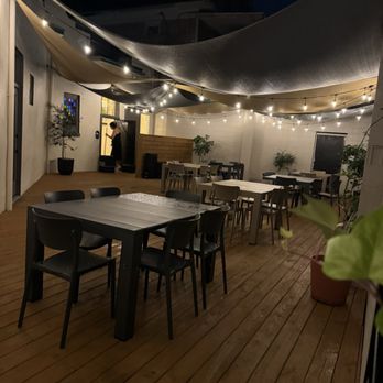 Othello - Outside patio - An outdoor patio area at night with wooden flooring, several tables and chairs, and string lights hanging overhead. Fabric canopies are stretched across the space, providing partial cover. Potted plants are placed along the walls, and a person is seen near the entrance or a doorway. The setting appears to be part of a restaurant or cafe, with ambient lighting creating a cozy atmosphere. - Seafood Near Me - Jacksonville, Florida