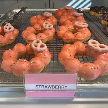 Mochinut-Gilbert - Strawberry donuts - A tray of strawberry donuts with pink glaze is arranged on a wire cooling rack. The donuts are decorated with multicolored sprinkles and small pretzels placed on top. The display is set in a bakery or cafe environment, with a sign labeled 'STRAWBERRY' in front of the tray. The background shows a kitchen counter with equipment and utensils. - Bubble Tea Near Me - Gilbert, Arizona