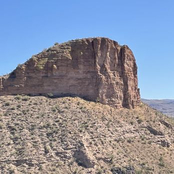 Apache Trail Tours - Desert scenery - A large sandstone rock formation dominates the scene in a desert environment, with a rugged, layered surface and sparse desert vegetation on the surrounding slopes. The sky is clear and blue, indicating a sunny day. The landscape appears arid with minimal plant life, typical of desert terrain. - Tours Near Me - Apache Junction, Arizona