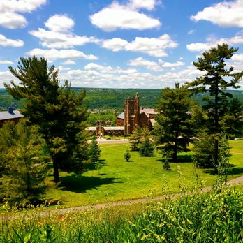 Cornell University - Cornell's lovely campus grounds - 5/29/2022 - A university campus scene with tall trees, green lawns, and a historic brick building, set against a backdrop of hills and a partly cloudy sky. - Colleges & Universities Near Me - Ithaca, New York
