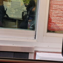 a woman sitting in a car - A person with dark hair is seen inside a pharmacy through a glass window, with a sign displaying COVID-19 testing instructions in Spanish.