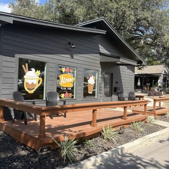 Atrium Coffee - A small coffee shop with gray siding and a wooden deck featuring black outdoor chairs. The shop has three windows with colorful signs, including images of coffee and ice cream, and the name 'Atrium' displayed. There is an 'Open' sign on the door, and a small 'No Parking' sign is visible in the landscaped area in front. The background includes trees and neighboring buildings, indicating a suburban setting. - Coffee & Tea Near Me - Belton, Texas