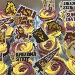 12 German chocolate and 12 funfetti cupcakes for our graduate's party. So yum for our GF/DF crew! - A collection of cupcakes with yellow and purple swirled frosting, decorated with various Arizona State University-themed toppers such as logos, mascots, and slogans. The cupcakes are arranged closely together, each in a metallic wrapper, and feature printed paper toppers representing ASU and Arizona State University.