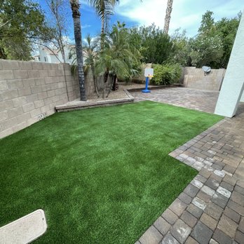 TheHardworkers - The image shows a backyard with artificial grass, brick pavers, a brick wall, and a small basketball hoop, with trees and shrubs in the background. - Landscape Architects or Designers Near Me - Mesa, Arizona