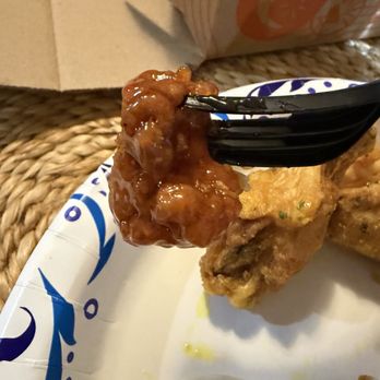 Wings Over Worcester - Considered one boneless piece - A piece of fried chicken with a sauce coating is held by a black fork above a paper plate with blue decorative patterns. The plate is placed on a woven surface, and part of a cardboard box is visible in the background. The image captures a close-up of the food and utensil, focusing on the fried chicken and sauce. - Chicken Wings Near Me - Worcester, Massachusetts