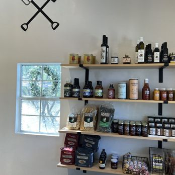 Steadfast Coffee - Store interior features wooden shelves stocked with pasta, sauces, and bottled products, positioned near a window that reveals trees outside. The wall above the shelves has a decal of a gardening fork and trowel crossed. The shelves hold various food items, including pasta, jars, and bottles, with some branded packaging visible. The lower part of the image shows hats and bags, and a wire basket is partially visible at the bottom left. - Coffee & Tea Near Me - Mesa, Arizona