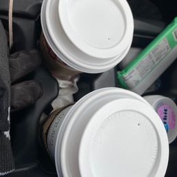 two cups of coffee in a cup holder - Two coffee cups with lids are held in a person's gloved hand, with a snack and other items visible in the background.