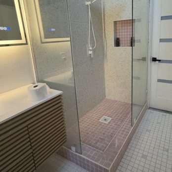 Bodega Style - Bathroom Tiles - The image shows a bathroom with a walk-in shower enclosed by glass walls. The shower has pink tiles on the floor and a small built-in niche with matching pink tiles. A white showerhead is mounted on the wall. To the left, there is a wooden cabinet with horizontal slats, and a paper towel roll is placed on top. A white door with horizontal black accents is visible on the right side of the image. - Building Supplies Near Me - Anaheim, California