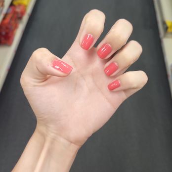 Charming Nails - A close-up of a person's hand with freshly painted coral-colored nails is shown, with the fingers slightly curled. The background features a store aisle with shelves stocked with snacks and other products, slightly out of focus. The lighting is bright, highlighting the glossy finish of the nail polish. - Nail Salons Near Me - Washington, DC, District of Columbia