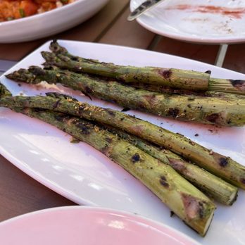 Steak + Vine - Asparagus - The image shows a white rectangular plate containing grilled asparagus spears with visible char marks, placed on a wooden table outdoors. In the background, there are other plates with food, indicating a dining setting, possibly at a restaurant or outdoor meal. - Steakhouses Near Me - La Grange, Illinois