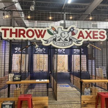 Celtic Axe Throwing - Austin - Indoor axe throwing lanes with six targets, safety barriers, and wooden dividers at Celtic Axe Throwing in Austin, TX. The area features a large sign reading 'THROW AXES' with a logo and flags, and is equipped with axes mounted on the wall. The space has industrial-style lighting and a concrete floor, with seating and tables nearby. - Axe Throwing Near Me - Austin, Texas