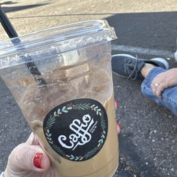 Iced coffee! - A hand with red-painted nails holds a clear plastic cup of iced coffee with a black circular logo that says 'Caffio' in stylized white text. The cup contains iced coffee with visible ice cubes and a black straw. The background shows a parking lot with several people walking and a person sitting nearby, wearing jeans and sneakers. The scene is outdoors with sunlight casting shadows.
