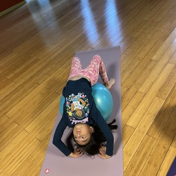 Barefoot Yoga Studio - A young girl is lying on her back on a yoga mat, with her head resting on her hands and a stability ball under her hips, in a room with wooden flooring and red curtains. - Yoga Near Me - Davis, California