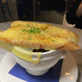 Le Sel - Onion soup - A bowl of French onion soup features a layer of melted cheese and toasted bread slices on top, garnished with chopped green onions. The soup is served in a white ceramic bowl placed on a white plate with a blue napkin underneath. The background shows a restaurant interior with dim lighting and wooden furniture, indicating a dining environment. - French Near Me - Washington, DC, District of Columbia