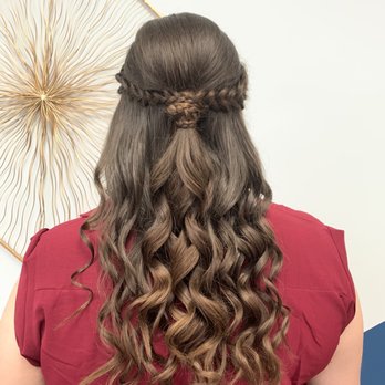 Meraki Salon - Formal style done 8/14/21 - A woman with long, brown, curly hair styled with a braided crown across the top of her head, with the rest of her hair flowing in loose curls down her back. She is wearing a red top and is photographed from behind, with a decorative wall art piece visible in the background. - Hair Salons Near Me - Antioch, Illinois