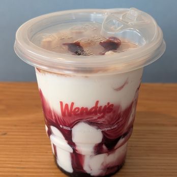 Wendy's - Cherry crumble vanilla frosty - A Wendy's cherry crumble vanilla Frosty in a clear plastic cup with a lid, topped with cherry pieces and syrup, placed on a wooden surface. - Burgers Near Me - Aurora, Ontario